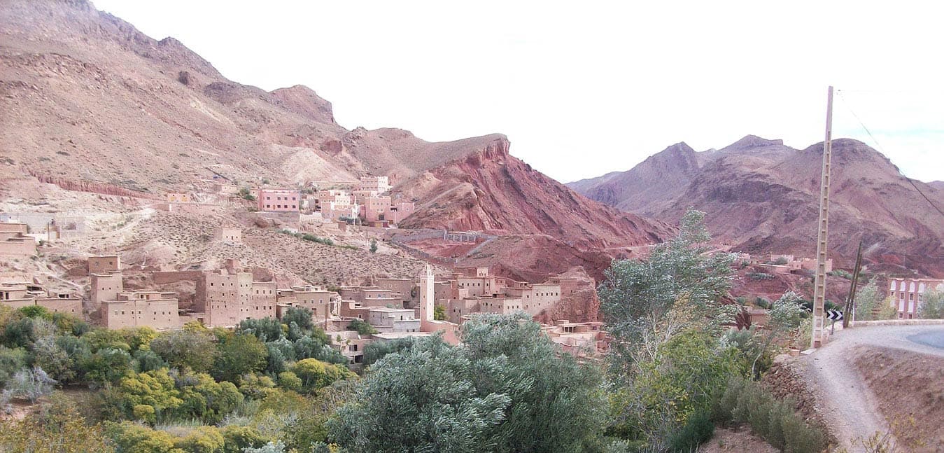 Earth-coloured village houses among green trees beneath red rocky mountains in Morocco