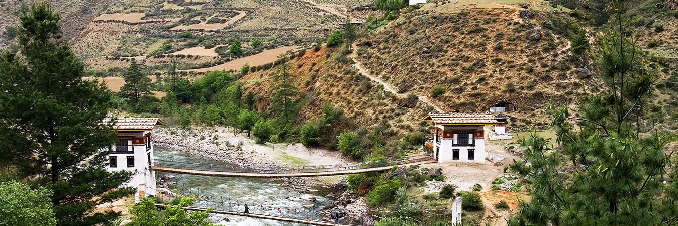 Chain footbridge with white gatehouses over a rocky river in a wooded Bhutanese valley