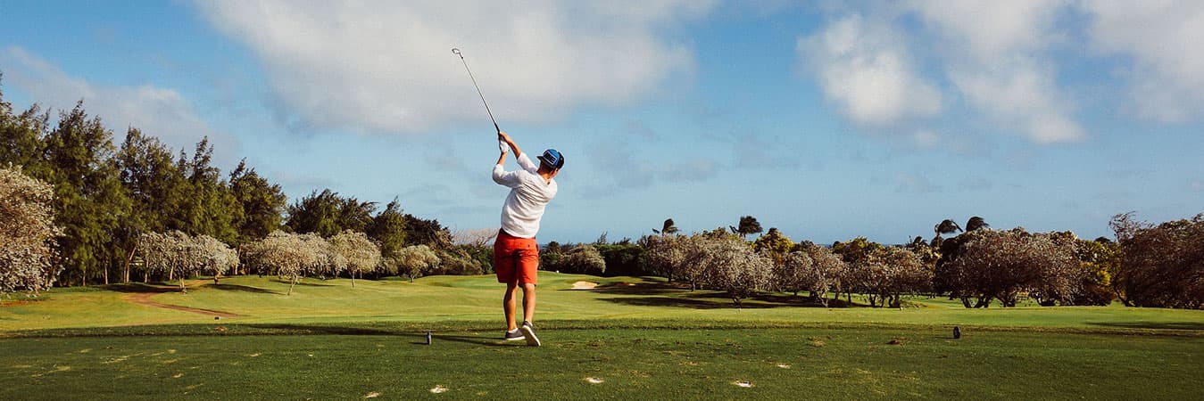 Golfer mid-swing on a green fairway lined with trees under a partly cloudy sky
