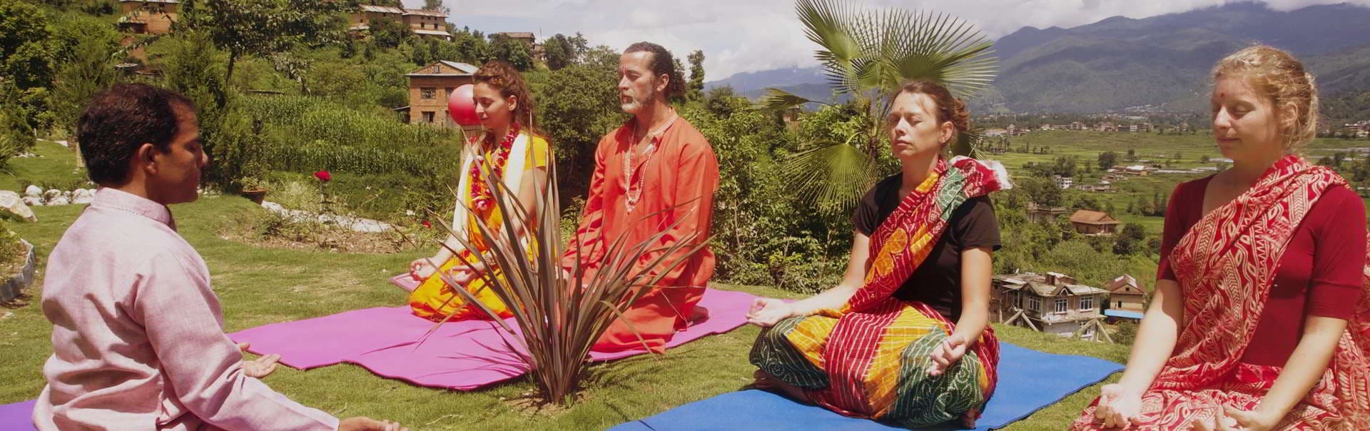 Group seated cross-legged on yoga mats on a lawn overlooking green hills and villages