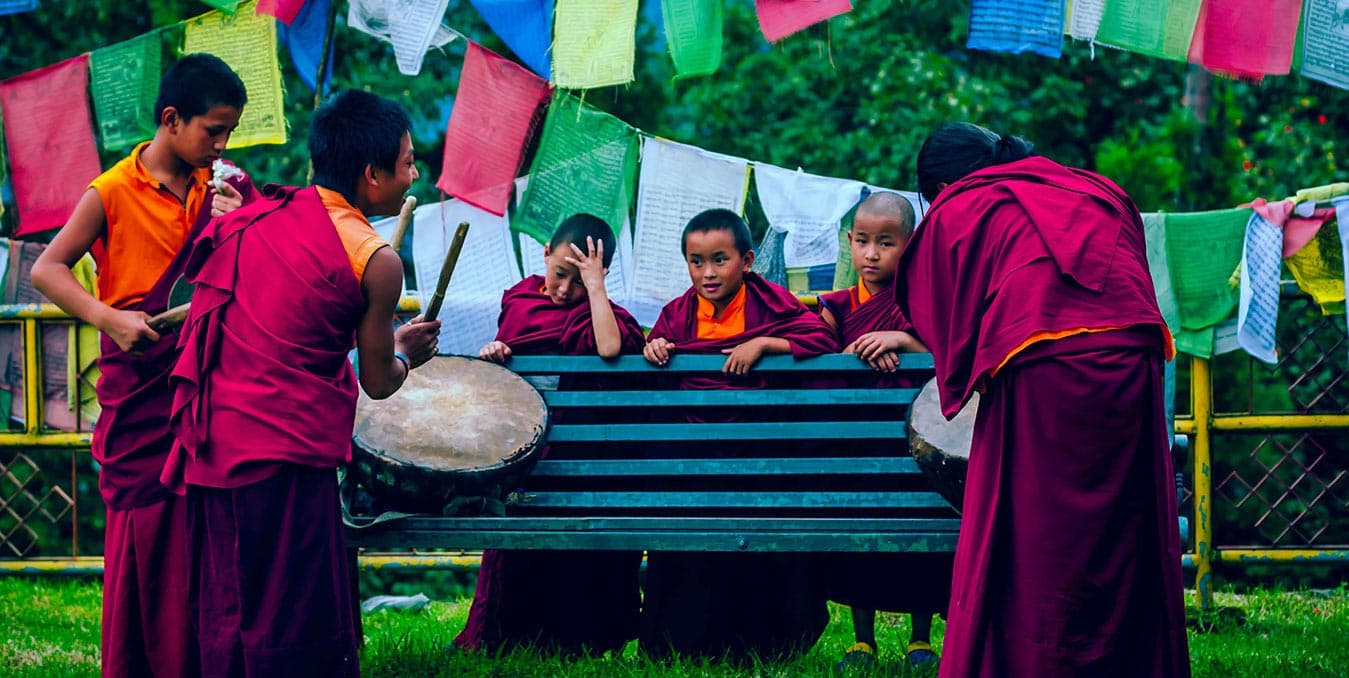 Young monks in maroon robes playing drums beside a bench under prayer flags