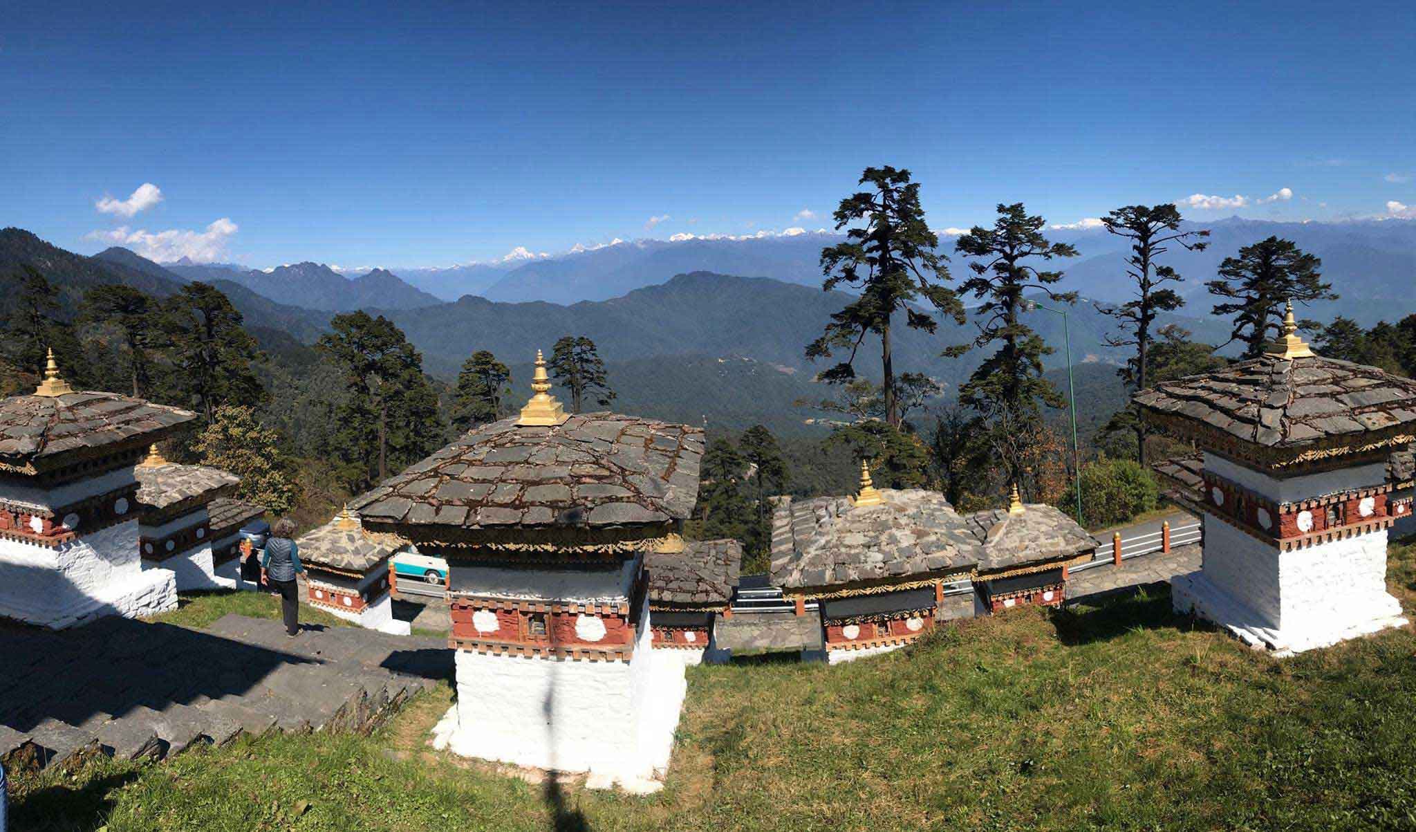Whitewashed chortens with slate roofs on a grassy pass overlooking forested hills in Bhutan