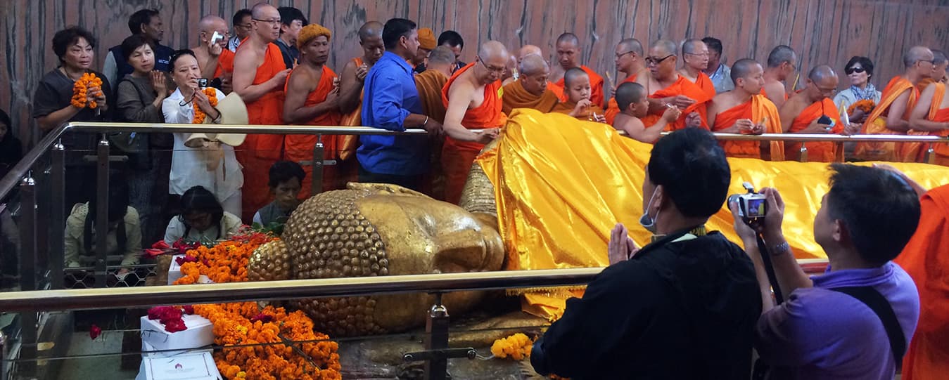 Monks in orange robes and pilgrims offering flowers beside a large golden reclining Buddha