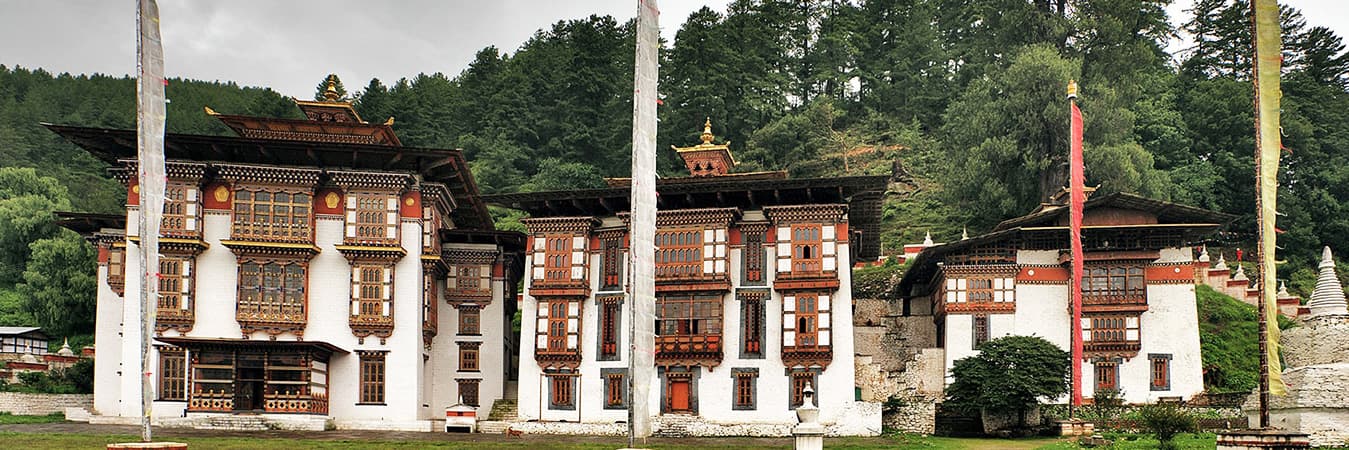 Whitewashed Bumthang temple buildings with prayer flag poles and a forested hillside behind