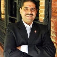 Rajan Simkhada smiling with arms folded, in a dark suit beside a brick wall