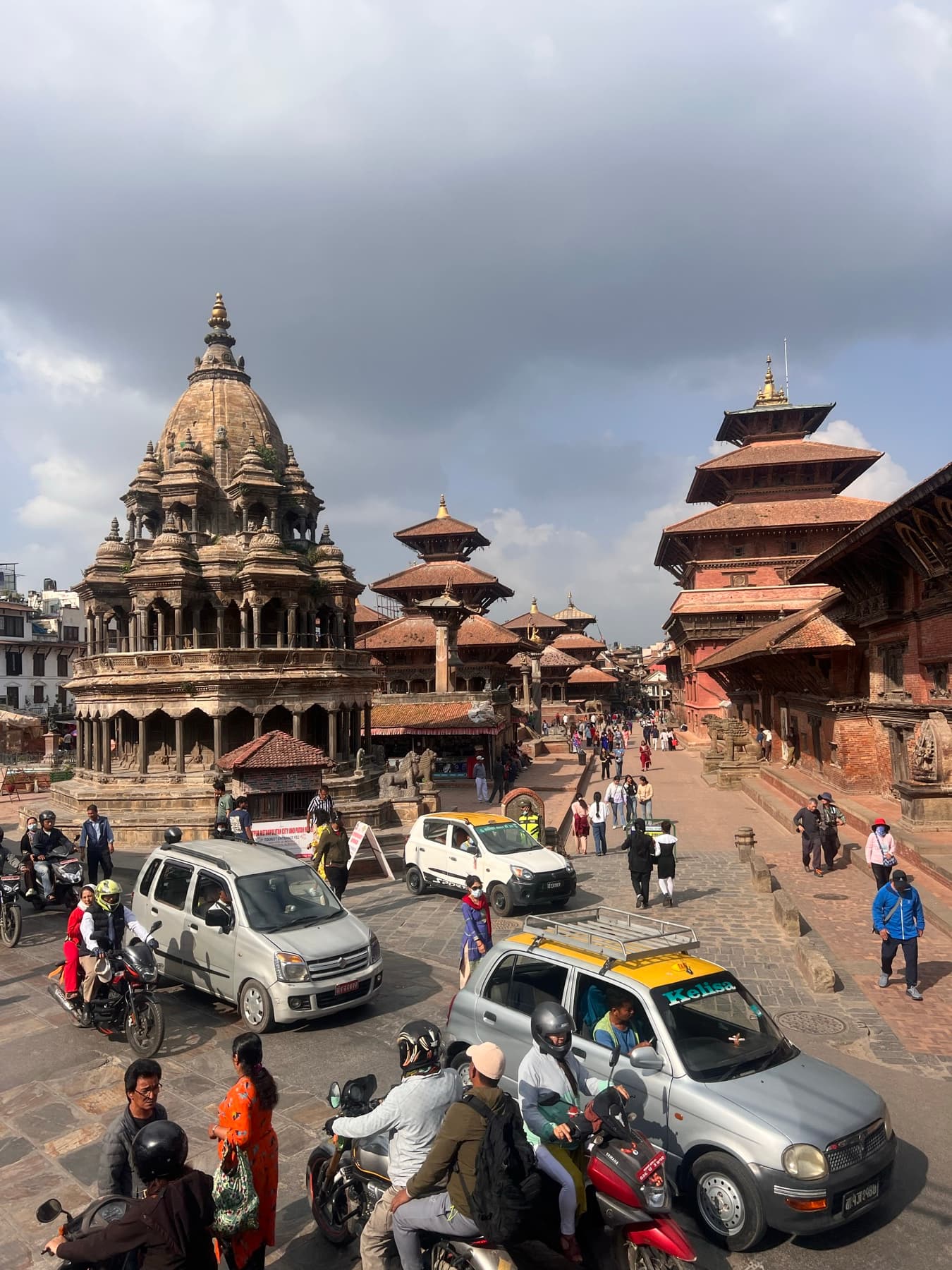 Temples and pagodas of a Durbar Square with cars, motorbikes and people in front