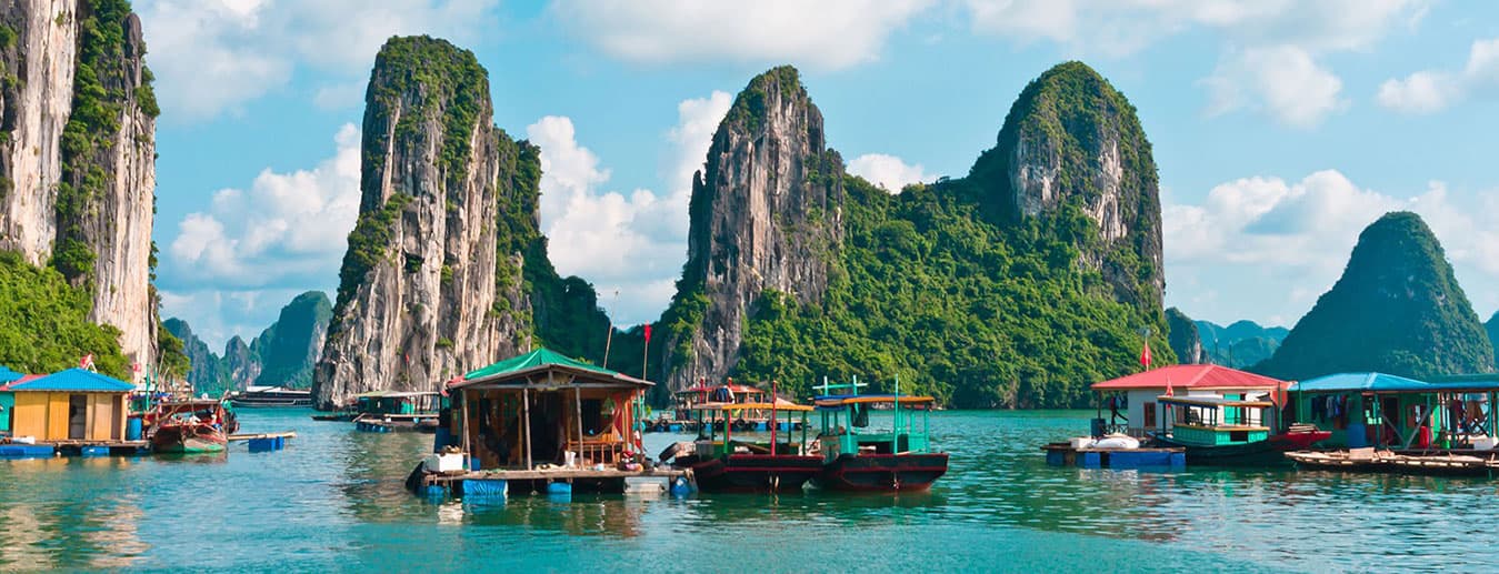 Floating houses and boats on calm water below tall forested limestone karst peaks