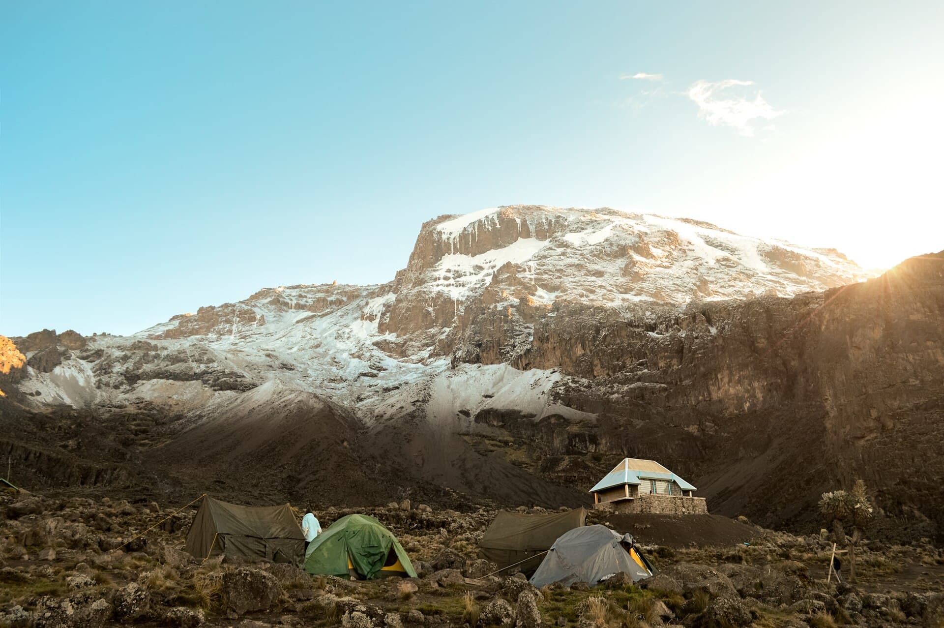 Tents and a small stone hut below the snow-streaked summit of Kilimanjaro at sunrise